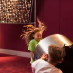 Zwei Kinder spielen zusammen im Haus; ein Kind schiebt einen großen zylindrischen Gegenstand, wodurch sich die Haare des anderen in die Luft heben.