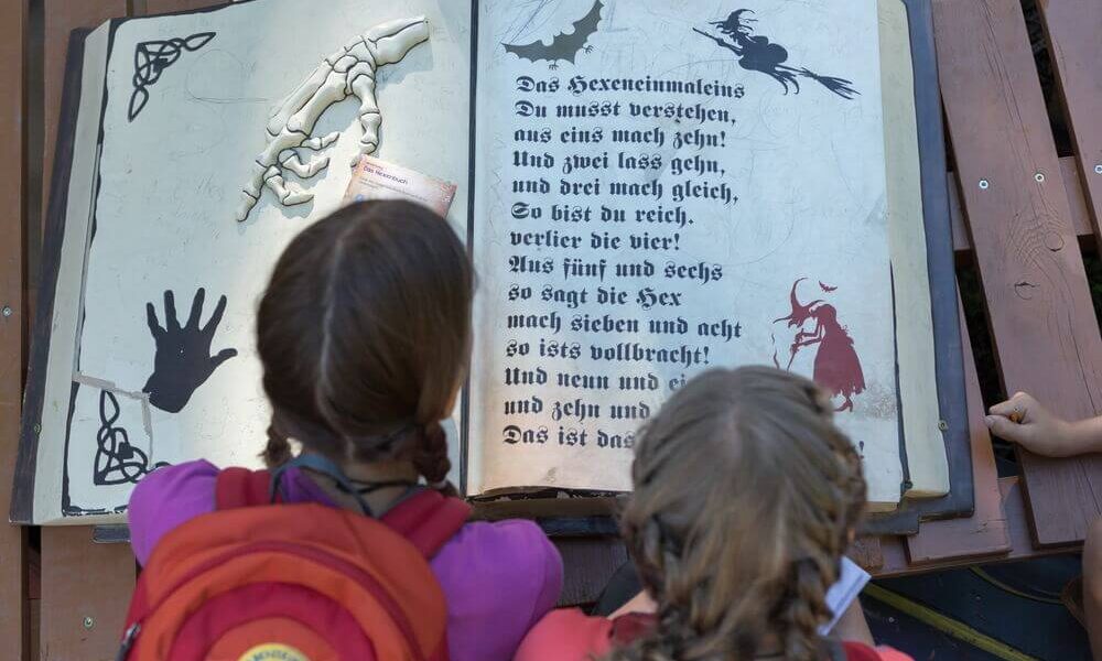Zwei Kinder mit Rucksäcken schauen auf ein großes aufgeschlagenes Buch mit deutschem Text, einer Skeletthand, einer Fledermaus und verschiedenen schwarzen Illustrationen auf den Seiten.