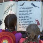 Zwei Kinder mit Rucksäcken schauen auf ein großes aufgeschlagenes Buch mit deutschem Text, einer Skeletthand, einer Fledermaus und verschiedenen schwarzen Illustrationen auf den Seiten.