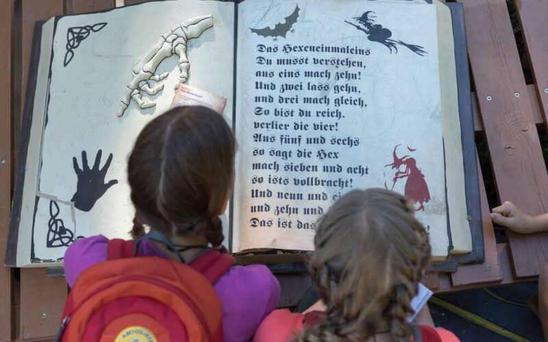 Zwei Kinder mit Rucksäcken schauen auf ein großes aufgeschlagenes Buch mit deutschem Text, einer Skeletthand, einer Fledermaus und verschiedenen schwarzen Illustrationen auf den Seiten.
