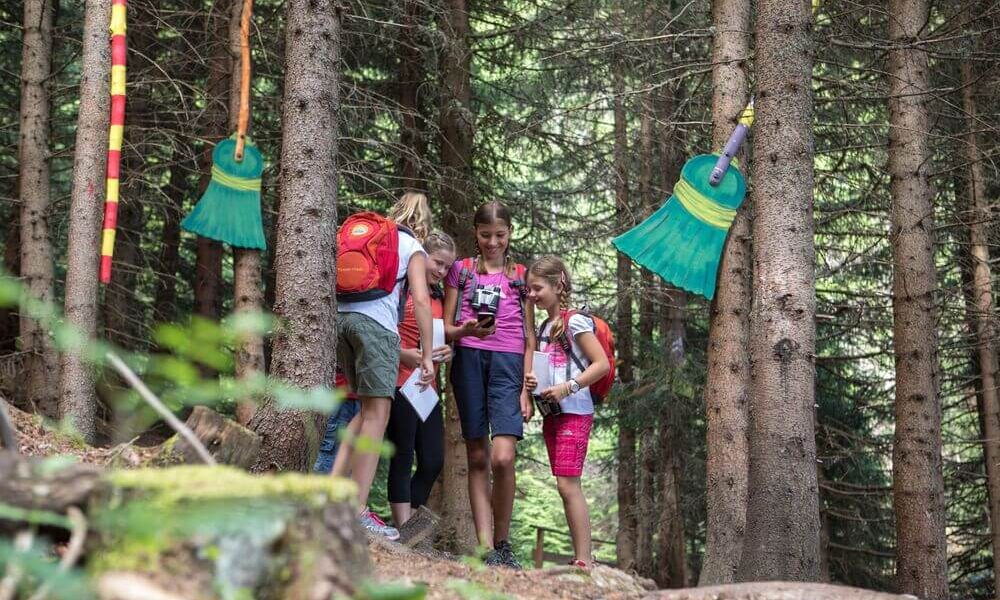 Vier Kinder mit Rucksäcken stehen zusammen in einem Wald und halten Papiere in der Hand, während in der Nähe große, bunte Besen an den Bäumen hängen.