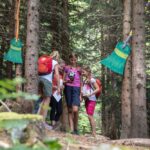 Vier Kinder mit Rucksäcken stehen zusammen in einem Wald und halten Papiere in der Hand, während in der Nähe große, bunte Besen an den Bäumen hängen.