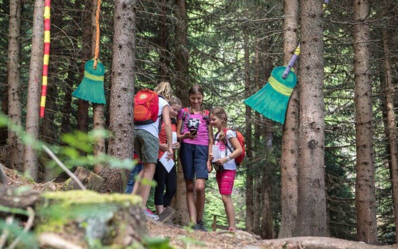 Vier Kinder mit Rucksäcken stehen zusammen in einem Wald und halten Papiere in der Hand, während in der Nähe große, bunte Besen an den Bäumen hängen.