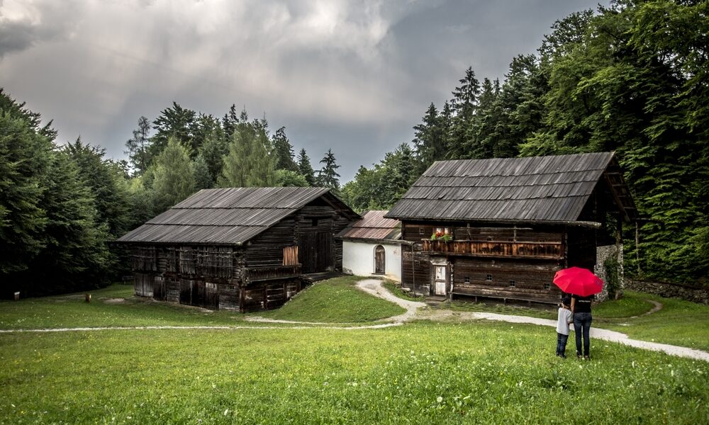 Zwei Personen mit einem roten Regenschirm stehen auf einer Wiese in der Nähe von zwei alten Holzhäusern, mit Bäumen und einem bewölkten Himmel im Hintergrund.