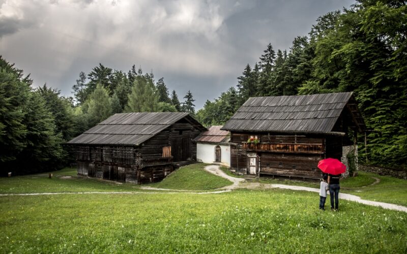 Zwei Personen mit einem roten Regenschirm stehen auf einer Wiese in der Nähe von zwei alten Holzhäusern, mit Bäumen und einem bewölkten Himmel im Hintergrund.