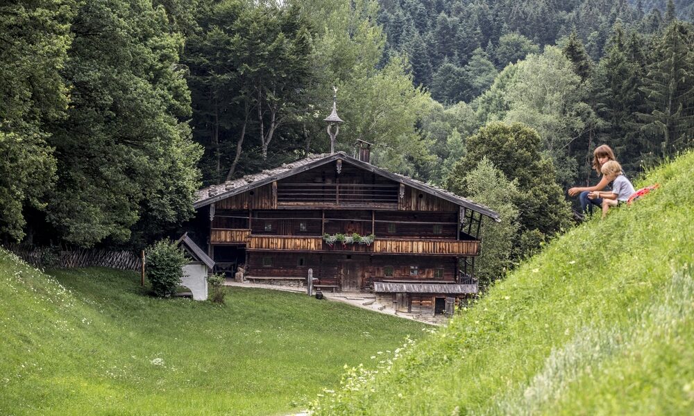 Eine rustikale Holzhütte steht zwischen Bäumen, im Vordergrund spielen ein Kind und ein Erwachsener auf einem grasbewachsenen Hügel.