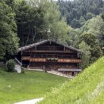 Eine rustikale Holzhütte steht zwischen Bäumen, im Vordergrund spielen ein Kind und ein Erwachsener auf einem grasbewachsenen Hügel.