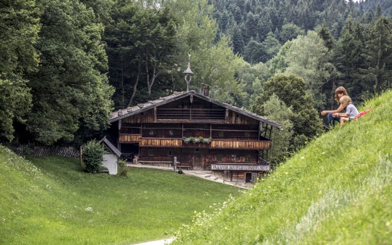Eine rustikale Holzhütte steht zwischen Bäumen, im Vordergrund spielen ein Kind und ein Erwachsener auf einem grasbewachsenen Hügel.