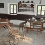 Altmodisches Zimmer mit Holzmöbeln, einem runden Tisch mit Tassen, Bänken, einem Rollstuhl aus Weidengeflecht, einem Schaukelpferd aus Holz, Wandregalen mit Geschirr und zwei kleinen Fenstern.