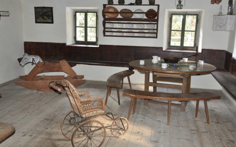 Altmodisches Zimmer mit Holzmöbeln, einem runden Tisch mit Tassen, Bänken, einem Rollstuhl aus Weidengeflecht, einem Schaukelpferd aus Holz, Wandregalen mit Geschirr und zwei kleinen Fenstern.