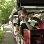 Zwei Jungen sitzen in einem offenen Zugwaggon, einer winkt, der andere zeigt, während der Zug an einem sonnigen Tag durch ein Waldgebiet fährt.