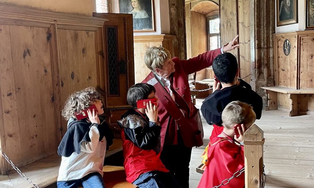 Eine Gruppe von Kindern und ein Erwachsener posieren spielerisch in einem historischen Raum mit Holzwänden, Porträts und gewölbten Türöffnungen.