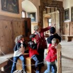 Eine Gruppe von Kindern und ein Erwachsener posieren spielerisch in einem historischen Raum mit Holzwänden, Porträts und gewölbten Türöffnungen.