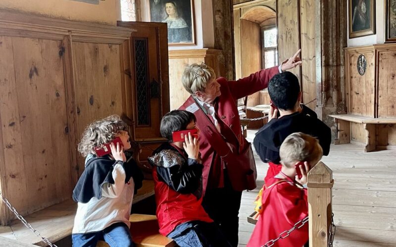 Eine Gruppe von Kindern und ein Erwachsener posieren spielerisch in einem historischen Raum mit Holzwänden, Porträts und gewölbten Türöffnungen.