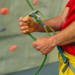 Eine Person, die einen Klettergurt trägt, benutzt ein Sicherungsgerät und ein grünes Kletterseil in einer Kletterhalle.