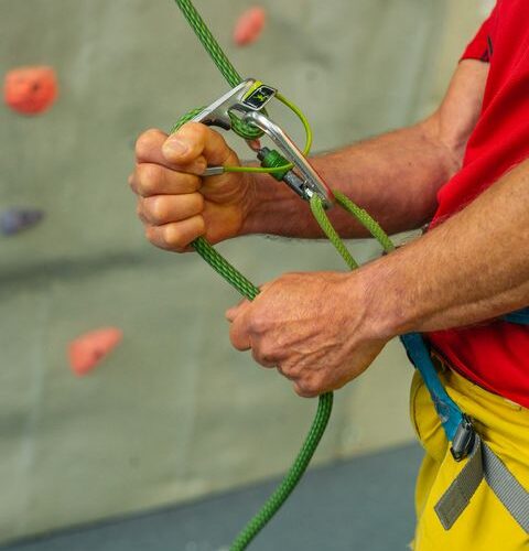 Eine Person, die einen Klettergurt trägt, benutzt ein Sicherungsgerät und ein grünes Kletterseil in einer Kletterhalle.