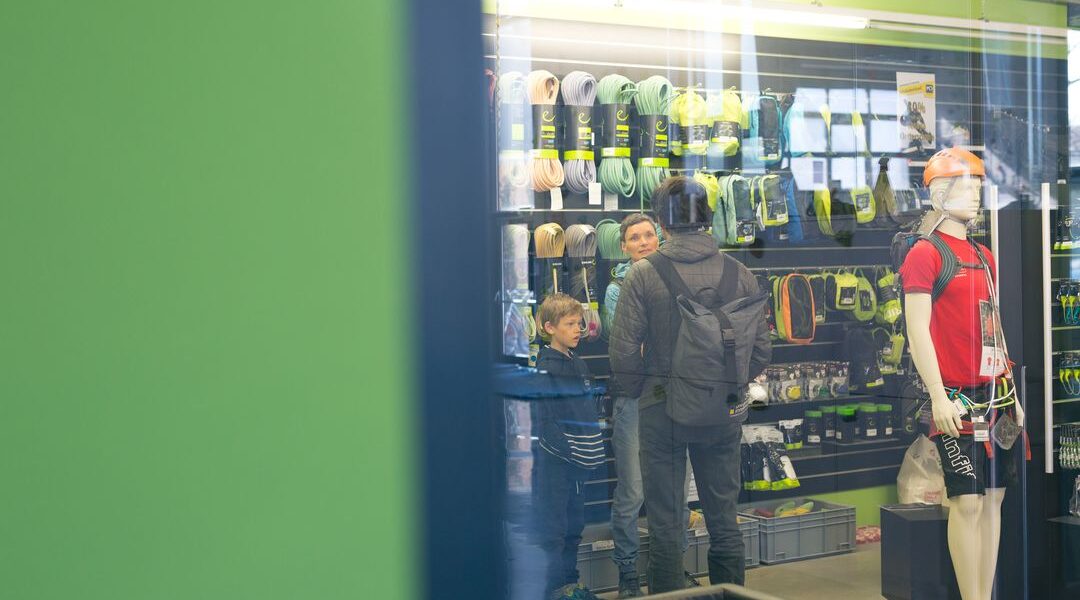 Kunden, darunter ein Mann und zwei Jungen, stöbern in einem Geschäft in Kletterausrüstung; eine Schaufensterpuppe mit Kletterausrüstung steht in der Nähe.