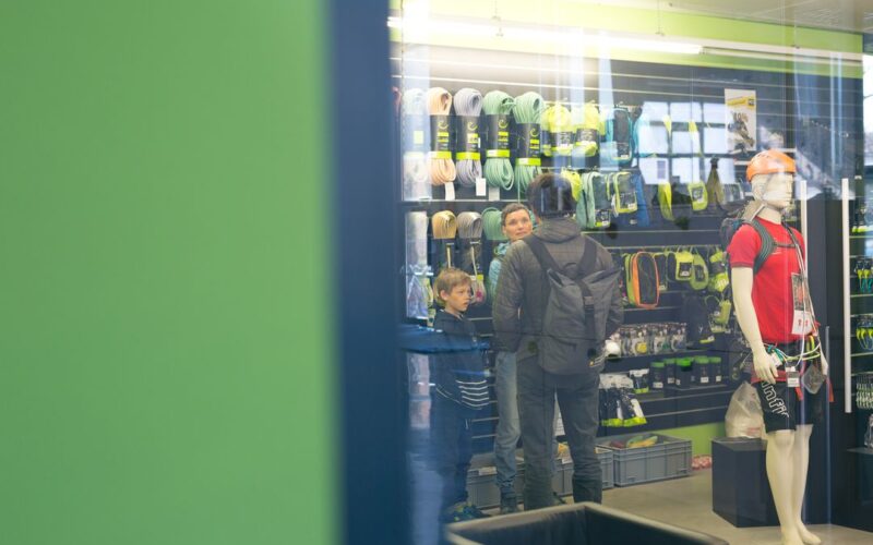Kunden, darunter ein Mann und zwei Jungen, stöbern in einem Geschäft in Kletterausrüstung; eine Schaufensterpuppe mit Kletterausrüstung steht in der Nähe.