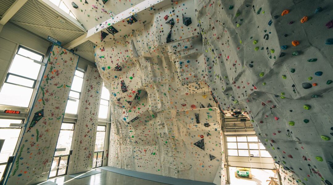Indoor-Kletterhalle mit hohen künstlichen Kletterwänden, bunten Griffen und poliertem Betonboden, beleuchtet von natürlichem Licht aus großen Fenstern.