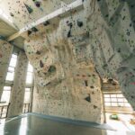 Indoor-Kletterhalle mit hohen künstlichen Kletterwänden, bunten Griffen und poliertem Betonboden, beleuchtet von natürlichem Licht aus großen Fenstern.