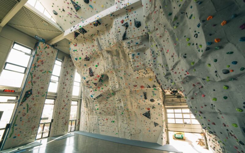 Indoor-Kletterhalle mit hohen künstlichen Kletterwänden, bunten Griffen und poliertem Betonboden, beleuchtet von natürlichem Licht aus großen Fenstern.