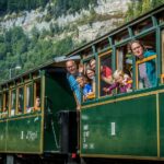 Fahrgäste, darunter Kinder und Erwachsene, lehnen sich aus den Fenstern und winken aus einem grünen Oldtimerzug, der durch eine bergige Gegend mit Wald im Hintergrund fährt.