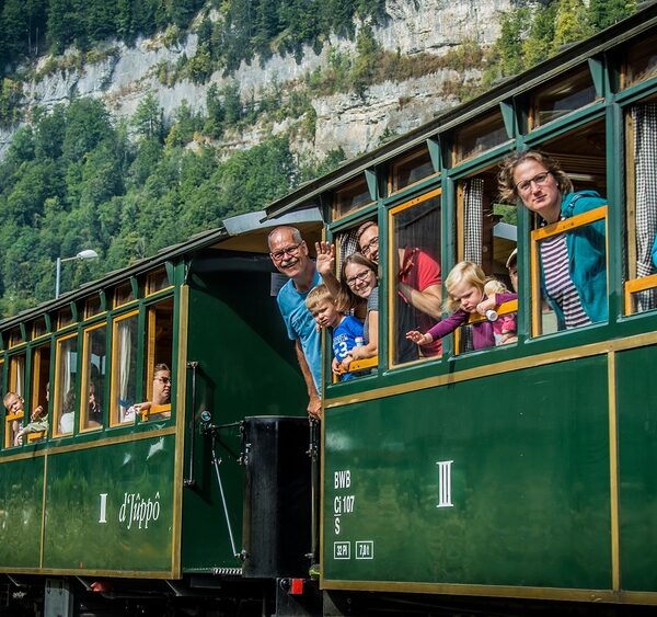 Fahrgäste, darunter Kinder und Erwachsene, lehnen sich aus den Fenstern und winken aus einem grünen Oldtimerzug, der durch eine bergige Gegend mit Wald im Hintergrund fährt.