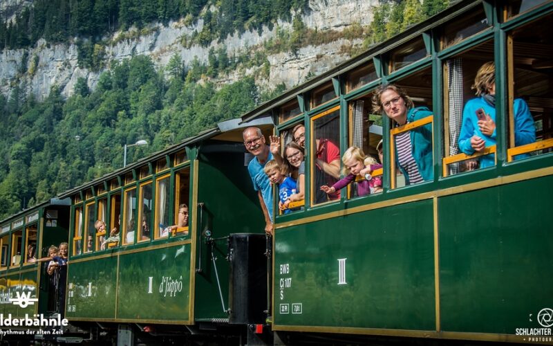 Fahrgäste, darunter Kinder und Erwachsene, lehnen sich aus den Fenstern und winken aus einem grünen Oldtimerzug, der durch eine bergige Gegend mit Wald im Hintergrund fährt.