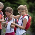 Drei Mädchen mit Rucksäcken und Ferngläsern stehen in einem Waldstück im Freien und schreiben und lesen aus Notizblöcken.