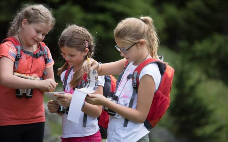 Drei Mädchen mit Rucksäcken und Ferngläsern stehen in einem Waldstück im Freien und schreiben und lesen aus Notizblöcken.