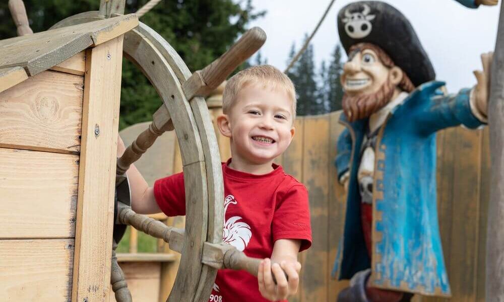 Ein Junge lächelt, während er das Rad eines hölzernen Spielgerüsts hält, mit einer großen Piratenstatue im Hintergrund.