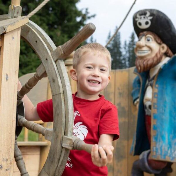 Ein Junge lächelt, während er das Rad eines hölzernen Spielgerüsts hält, mit einer großen Piratenstatue im Hintergrund.