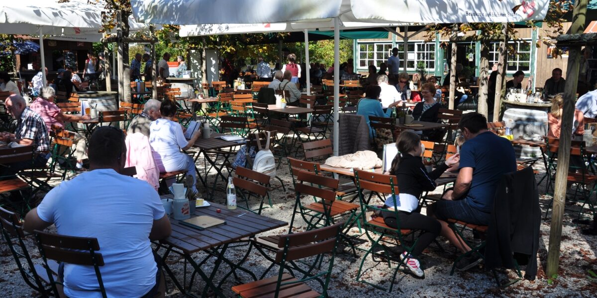 An einem sonnigen Tag sitzen die Menschen an Holztischen unter großen weißen Sonnenschirmen in einem Café oder Biergarten im Freien, umgeben von Bäumen und Kiesboden.