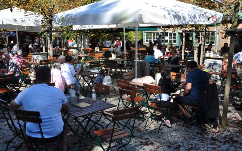 An einem sonnigen Tag sitzen die Menschen an Holztischen unter großen weißen Sonnenschirmen in einem Café oder Biergarten im Freien, umgeben von Bäumen und Kiesboden.
