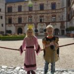 Zwei Kinder in mittelalterlichen Kostümen, eines als Prinzessin und eines als Ritter, stehen draußen vor einem historischen Gebäude.