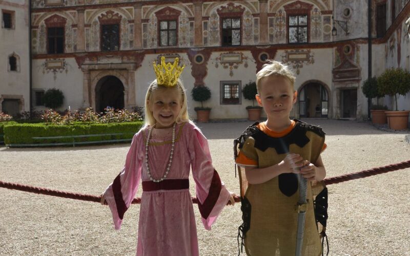 Zwei Kinder in mittelalterlichen Kostümen, eines als Prinzessin und eines als Ritter, stehen draußen vor einem historischen Gebäude.