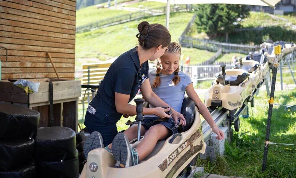 Eine Frau hilft einem jungen Mädchen, das in einem Bob-ähnlichen Wagen sitzt, bei einer Bahnfahrt und bereitet es an einem sonnigen Tag auf die Aktivität vor.