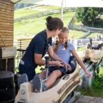 Eine Frau hilft einem jungen Mädchen, das in einem Bob-ähnlichen Wagen sitzt, bei einer Bahnfahrt und bereitet es an einem sonnigen Tag auf die Aktivität vor.