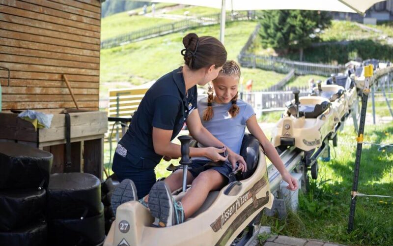 Eine Frau hilft einem jungen Mädchen, das in einem Bob-ähnlichen Wagen sitzt, bei einer Bahnfahrt und bereitet es an einem sonnigen Tag auf die Aktivität vor.