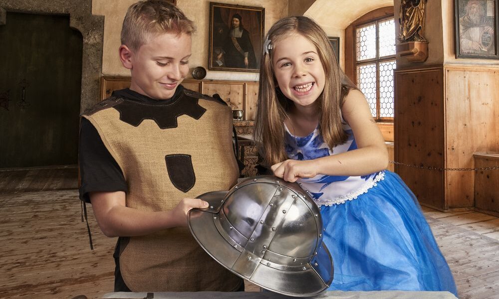 Zwei Kinder in Kostümen untersuchen einen mittelalterlichen Helm und ein Schwert in einem holzgetäfelten historischen Raum mit Gemälden an den Wänden.
