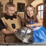 Zwei Kinder in Kostümen untersuchen einen mittelalterlichen Helm und ein Schwert in einem holzgetäfelten historischen Raum mit Gemälden an den Wänden.