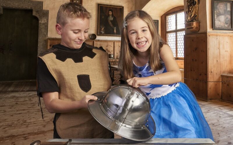 Zwei Kinder in Kostümen untersuchen einen mittelalterlichen Helm und ein Schwert in einem holzgetäfelten historischen Raum mit Gemälden an den Wänden.