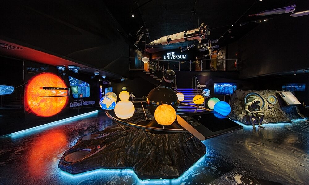 Eine wissenschaftliche Ausstellung in einem Innenraum zeigt beleuchtete Modelle des Sonnensystems, eine Raketennachbildung und Lehrtafeln zur Weltraumforschung.