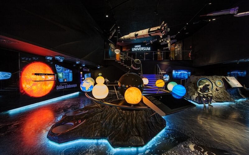 Eine wissenschaftliche Ausstellung in einem Innenraum zeigt beleuchtete Modelle des Sonnensystems, eine Raketennachbildung und Lehrtafeln zur Weltraumforschung.