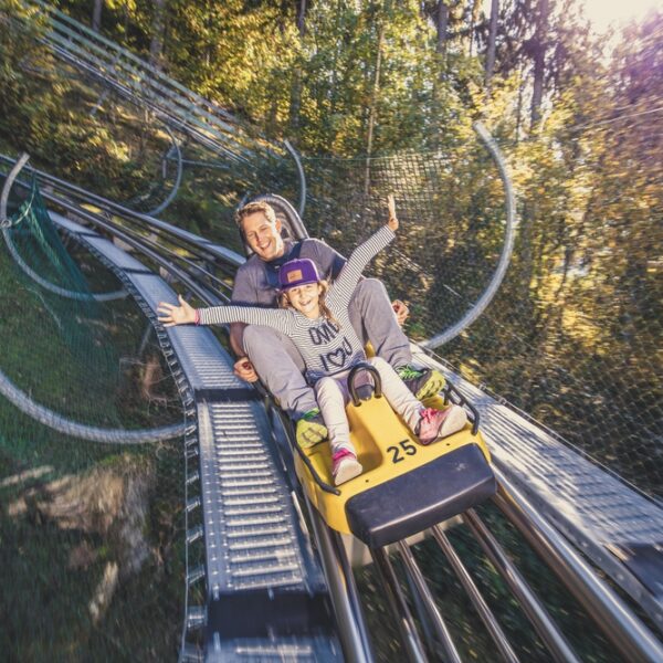 Ein Erwachsener und ein Kind fahren gemeinsam auf einem gelben Alpine Coaster durch ein bewaldetes Gebiet, beide lächeln mit erhobenen Armen.