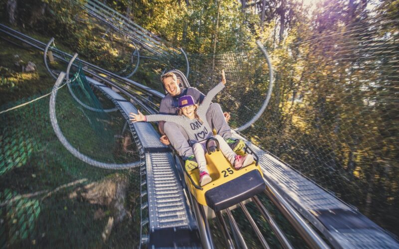 Ein Erwachsener und ein Kind fahren gemeinsam auf einem gelben Alpine Coaster durch ein bewaldetes Gebiet, beide lächeln mit erhobenen Armen.