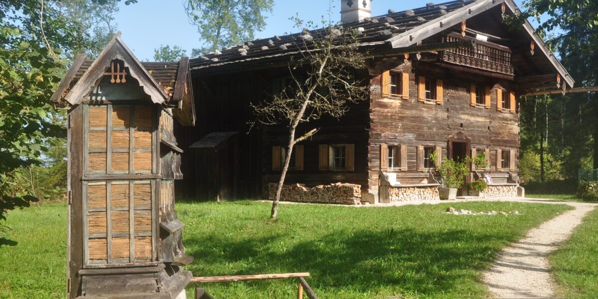 Ein rustikales Holzhaus mit Ziegeldach steht in einem grasbewachsenen Hof, mit einer traditionellen Holzkonstruktion und einem Feldweg im Vordergrund.