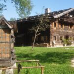 Ein rustikales Holzhaus mit Ziegeldach steht in einem grasbewachsenen Hof, mit einer traditionellen Holzkonstruktion und einem Feldweg im Vordergrund.