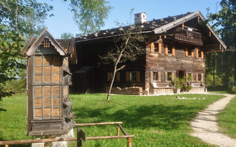Ein rustikales Holzhaus mit Ziegeldach steht in einem grasbewachsenen Hof, mit einer traditionellen Holzkonstruktion und einem Feldweg im Vordergrund.