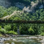 Ein Dampfzug überquert eine grüne Metallbrücke über einen felsigen Fluss, der von dichten grünen Wäldern und steilen Klippen umgeben ist.